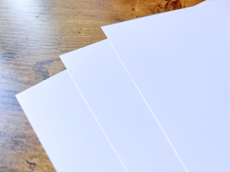 K&ouml;nnte beinhalten: Drei Bl&auml;tter wei&szlig;es Papier sind aufgef&auml;chert, ihre Kanten sind auf einer Holzoberfl&auml;che sichtbar. Das Papier ist strahlend wei&szlig; und das Holz hat einen warmen, braunen Ton. Die Komposition deutet auf Schreibwaren oder Kunstbedarf hin.