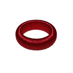Puede incluir: Un brazalete de seda rojo. El brazalete está hecho de un cordón de seda grueso y tejido y tiene un diseño simple y elegante.