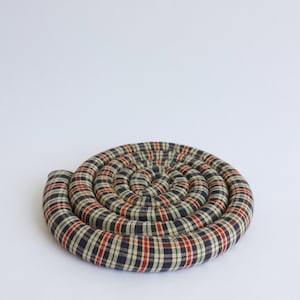 May include: A circular, coiled trivet with a plaid pattern in navy, red, green, and beige. The trivet is made of fabric and is designed to protect surfaces from hot items. It has a spiral shape.