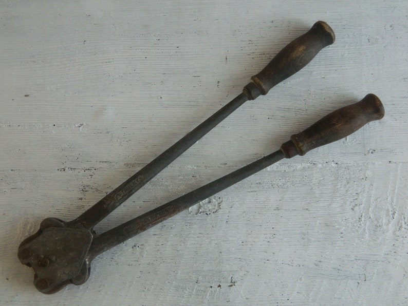 K&ouml;nnte beinhalten: Ein Vintage-Werkzeug aus Metall mit zwei Holzgriffen. Das Werkzeug hat einen Metallkopf mit einem Hebel und eine Metallplatte mit einem Loch in der Mitte. Das Werkzeug wird zum Verpressen von Metall verwendet.