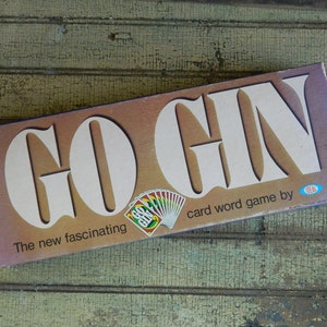 May include: A vintage box game called "Go Gin" with a brown background and white lettering. The box features a fan of playing cards with the words "Go Gin" on them. The text on the box reads "The new fascinating card word game by Ideal."