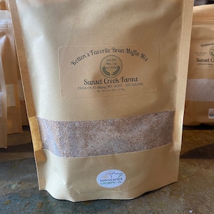May include: A brown paper bag with a white label that reads "Kotten's Favorite Bran Muffin Mix" and "Sunset Creek Farms". The bag contains a light brown powder. The label also includes the text "Just Add Milk, Egg and Butter" and "24378 CR 117 Albany, MN 56307 320.333.2140". The bag has a sticker that says "Minnesota Grown".