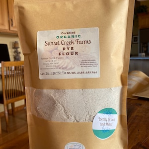 May include: A brown paper bag with a white label that reads "Certified Organic Sunset Creek Farms Rye Flour". The bag contains a white powder and has a sticker that says "Locally Grown and Milled".