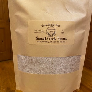 May include: A brown paper bag with a clear window showing a brown bran muffin mix. The bag has a label that reads "Bran Muffin Mix" and "Sunset Creek Farms". The label also says "Made with Organic Wheat Flour and Bran" and "Just add water, 1 egg, oil, honey".