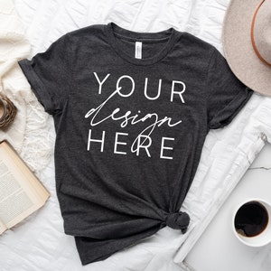 May include: A dark gray t-shirt with the text "YOUR design HERE" in white. The shirt is laid flat on a white surface with a book, a candle, a hat, and a cup of coffee.