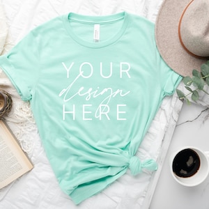May include: A light turquoise t-shirt with a white design that says "YOUR design HERE" on a white background. The t-shirt is knotted at the bottom and is lying on a white surface with a book, a clock, a cup of coffee, a hat, and a tray.