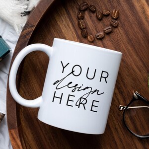 May include: White ceramic mug with a black inscription that reads "YOUR design HERE" on a wooden tray with coffee beans, a potted plant, and a pair of glasses.