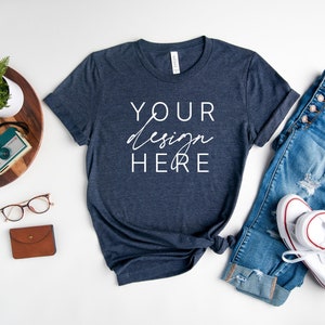 May include: A navy blue t-shirt with the text "YOUR design HERE" in white. The t-shirt is laid flat on a white surface with a pair of blue jeans, white sneakers, a wooden tray with a plant, a watch, and a camera.