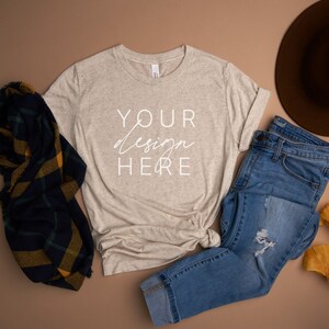 Pode incluir: Uma camiseta bege com o texto "YOUR design HERE" é combinada com jeans azuis, um chapéu marrom e um cachecol xadrez. Folhas amarelas adicionam um toque de outono. Os itens são dispostos em uma superfície marrom.