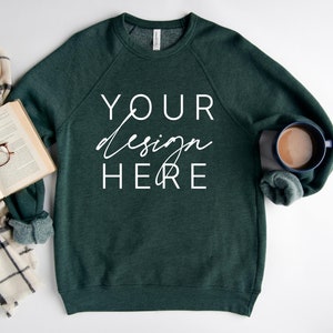以下が含まれることがあります： 白い背景に置かれた、濃い緑色のスウェットシャツ。前面に白い文字で「YOUR design HERE」と書いてある。スウェットシャツの隣には、本、眼鏡、スカーフが置かれている。