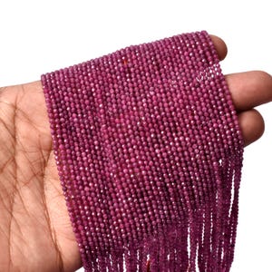 May include: A long strand of small, faceted, deep magenta-coloured beads arranged in multiple parallel rows. The beads have a uniform, circular shape with a reflective, textured surface that catches the light, creating a consistent, vibrant appearance.