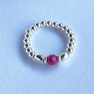 May include: A silver beaded ring featuring a faceted, deep red gemstone. The ring is made up of small, round beads and elongated silver elements that frame the gemstone. The background is a plain white surface.