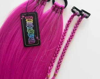 May include: Two bright pink hair extensions attached to black elastic hair ties, featuring a black rectangular tag with the text BRAIDAPALOOZA in colourful letters, alongside two smaller braided pink hair extensions with metal clips.