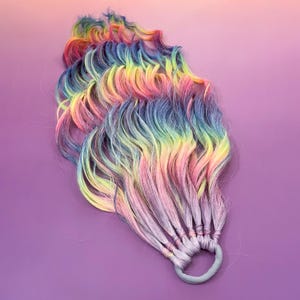 Puede incluir: Extensiones de cabello de color arcoíris con una banda elástica gris claro. Las extensiones presentan una mezcla de colores vibrantes, incluyendo rosa, amarillo, azul y verde, creando un efecto multicolor. Las hebras onduladas están agrupadas en la base, ofreciendo un accesorio lúdico y colorido.