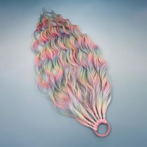 Puede incluir: Extensiones de cabello de color arcoíris pastel con textura ondulada. El cabello es una mezcla de tonos rosa, amarillo, azul y verde, sujeto a una goma para el pelo rosa. Las extensiones están diseñadas para ser voluminosas y fluidas.
