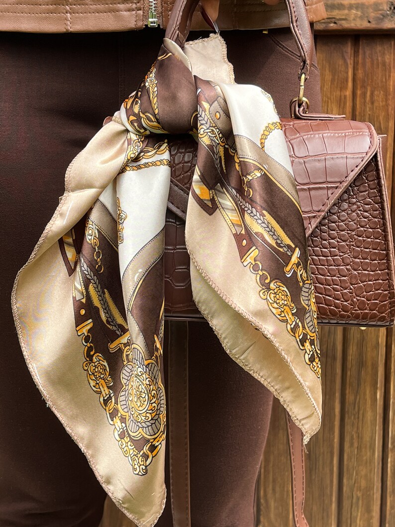 May include: A brown and beige patterned silk scarf tied around a brown leather handbag with a crocodile embossed design.
