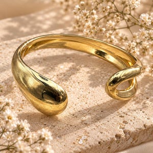May include: A polished gold-tone open cuff bracelet featuring a smooth, rounded, teardrop-shaped terminal on one end and a curved, tapered design on the other. The metal surface has a reflective, high-shine finish with a continuous, fluid shape.