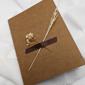 May include: A brown card with a piece of brown tape and dried flowers on top. The card is on a white background.