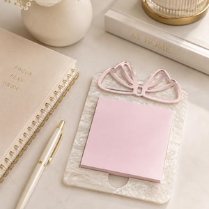 Op de afbeelding: Een rechthoekige memoblokhouder met een witte marmeren afwerking en een metallic roze strikaccent aan de bovenkant. De houder bevat een stapel vierkante lichtroze memoblaadjes. Het ontwerp heeft een glad, gepolijst oppervlak met een uitsparing.