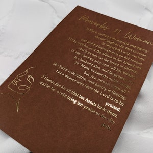 May include: A brown card with gold lettering featuring a quote from Proverbs 31 about a woman's strength, dignity, and wisdom. The quote reads, "She is clothed with strength and dignity; she can laugh at the days to come. She watches over the affairs of her household and faithful instruction is on her tongue. She speaks with wisdom. Her children arise and call her blessed; her husband also, and he praises her. Many women do noble things, but you surpass them all." The card also features a gold line drawing of a woman's face.