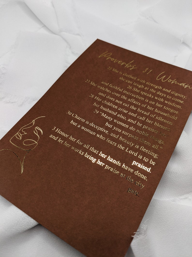 May include: A brown card with gold lettering featuring a quote from Proverbs 31 about a woman's strength, dignity, and wisdom. The quote reads, "She is clothed with strength and dignity; she can laugh at the days to come. She watches over the affairs of her household and faithful instruction is on her tongue. She speaks with wisdom. Her children arise and call her blessed; her husband also, and he praises her. Many women do noble things, but you surpass them all." The card also features a gold line drawing of a woman's face.