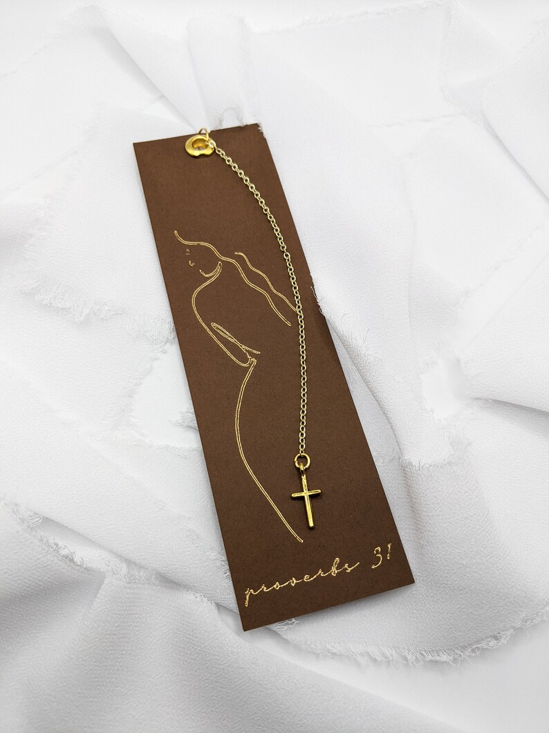 May include: A brown bookmark with a gold chain and a small gold cross charm. The bookmark has a gold outline of a woman and the text "proverbs 3!"