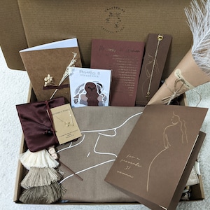 May include: A brown gift box filled with various items, including a brown card with the text "For a prayerful woman", a sticker pack with the text "Proverbs 31 Sticker Pack", a brown pouch with a gold chain necklace, a brown card with the text "Proverbs 31 Woman", a white feather bouquet, and a brown notebook.