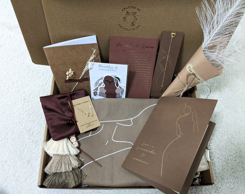 May include: A brown gift box filled with various items, including a brown card with the text "For a prayerful woman", a sticker pack with the text "Proverbs 31 Sticker Pack", a brown pouch with a gold chain necklace, a brown card with the text "Proverbs 31 Woman", a white feather bouquet, and a brown notebook.