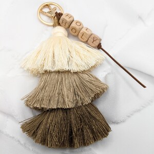 May include: A beige, brown, and dark brown tassel keychain with a gold key ring and a wooden bead charm that says "HOLY".