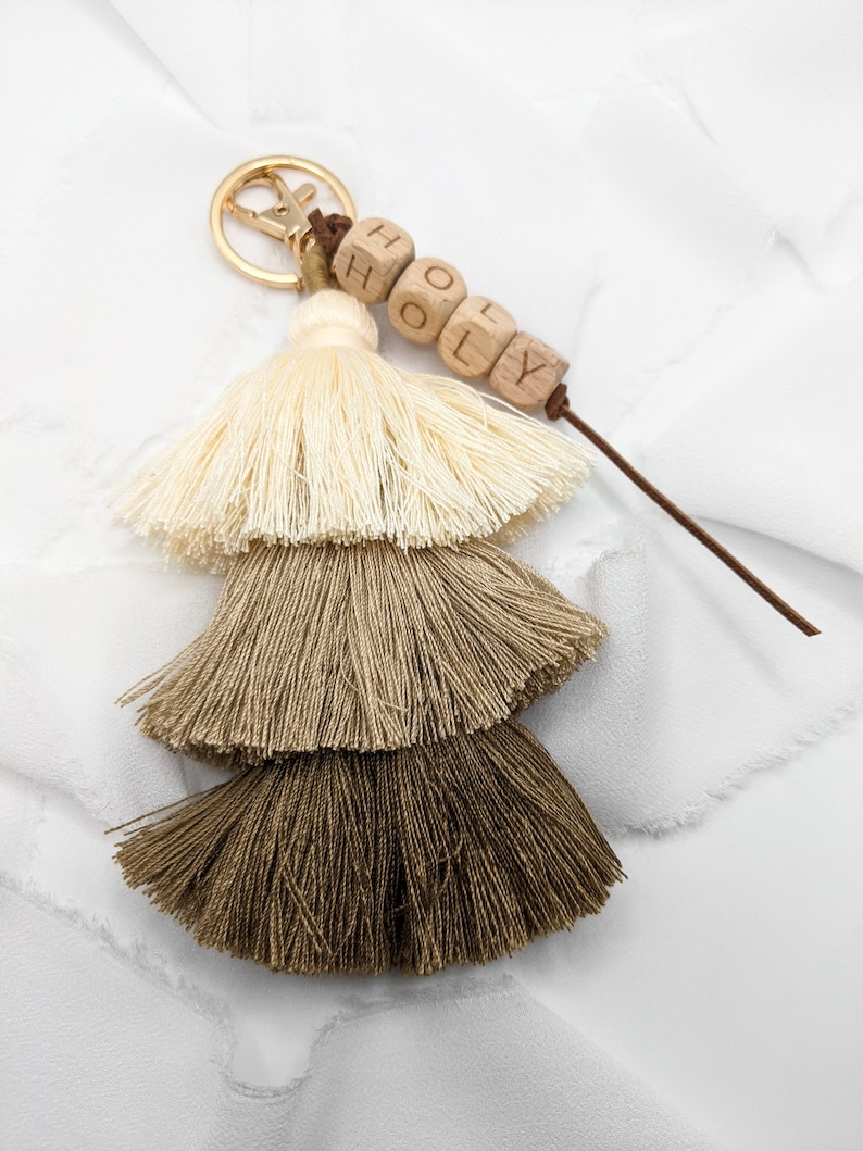 May include: A beige, brown, and dark brown tassel keychain with a gold key ring and a wooden bead charm that says "HOLY".