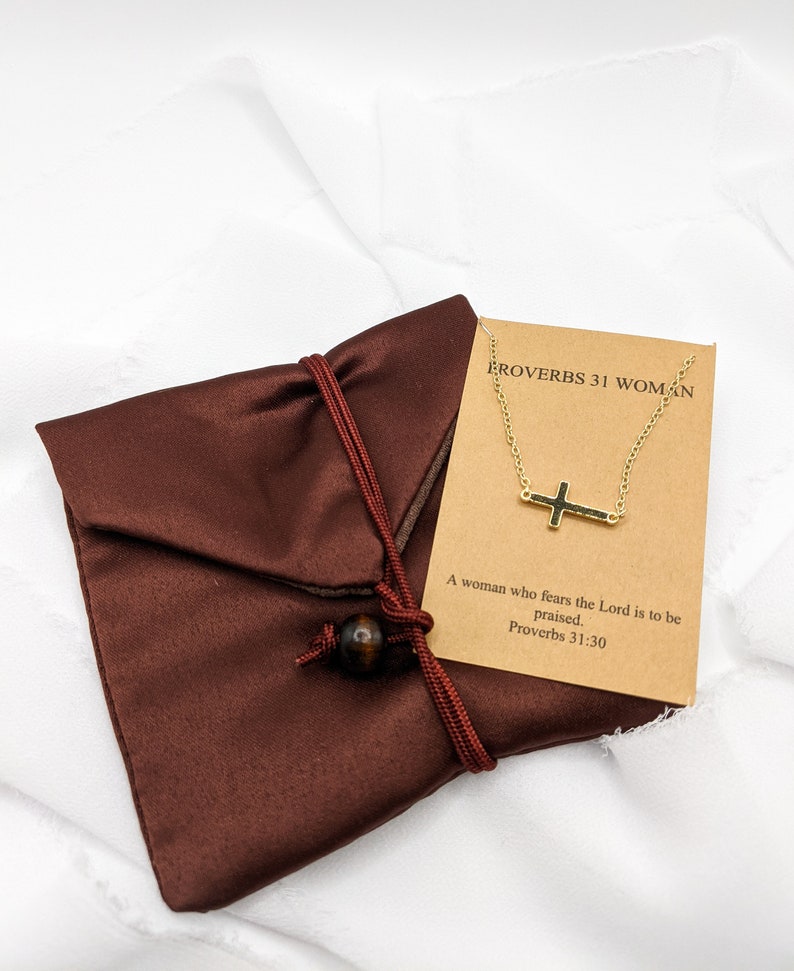 May include: A brown velvet pouch with a gold chain necklace featuring a small cross pendant. The card attached to the necklace reads "Proverbs 31 Woman" and "A woman who fears the Lord is to be praised. Proverbs 31:30".