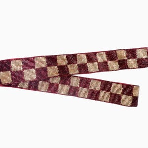 May include: A beaded purse strap with a burgundy and gold checkerboard pattern. The strap has a red backing and gold-colored metal clasps. The strap is designed to be attached to a purse or bag.