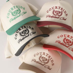 May include: Five white baseball caps with black and red embroidery. The caps have a black or red brim and feature the text "COURTSIDE Pickleball Club" with a logo of a pickleball and a wreath.