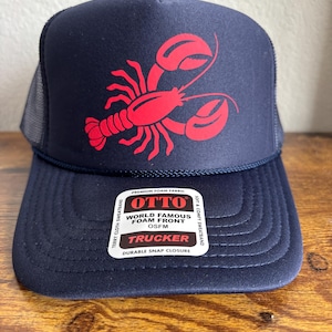 Puede incluir: Gorra de camionero azul marino con un diseño de langosta roja. La gorra tiene una parte trasera de malla y una visera curva. La parte delantera de la gorra tiene el texto "OTTO WORLD FAMOUS FOAM FRONT TRUCKER". Tiene un cierre a presión duradero.