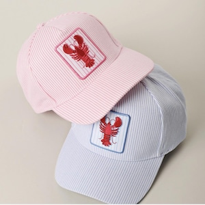 May include: Two baseball caps with a seersucker stripe pattern. One is pink, the other is light blue. Each cap features a red lobster patch with a white and blue border. The caps have a curved brim.