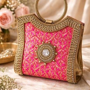 May include: A hot pink clutch with a gold handle and trim. The clutch features an intricate floral pattern and is embellished with gold beads and a large, central crystal. The handle is a circular gold ring.