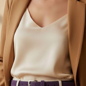 May include: A tan blazer over a cream-colored V-neck top, paired with cream-colored pants and a purple belt with a silver buckle. The outfit is a neutral color palette.
