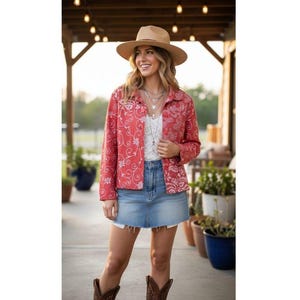 May include: A woman wearing a red jacket with a white floral pattern, a white top, and a denim skirt. She is also wearing a brown hat and boots. The background includes plants and a wooden structure.