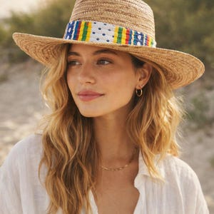 May include: A straw fedora hat with a wide brim. The hat features a colorful band with blue, yellow, green, and red stripes, and white polka dots. The hat is worn by a person with long, wavy blonde hair.