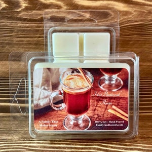 May include: A clear plastic container with four wax melts inside. The wax melts are scented with Hot Buttered Rum. The label features a photo of a mug of hot buttered rum with cinnamon sticks and the text "Hot Buttered Rum", "A Family That Makes Scents", "100% Soy - Hand-Poured", and "Familycandlescents.com".
