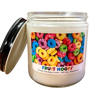 May include: A white jar candle with a black lid, featuring a label with colorful cereal rings. The label reads "FRUIT HOOPS" and "A Family That Makes Scents - 100% Soy - Hand-Poured". The lid is off to the side.