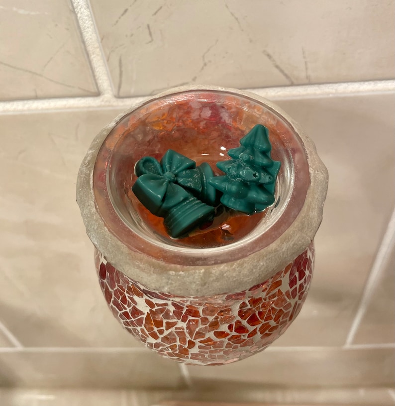 Pode incluir: Um aquecedor de cera de vidro com mosaico vermelho e branco com um prato de vidro transparente que cont&eacute;m tr&ecirc;s derretimentos de cera verde em forma de &aacute;rvore de Natal e um la&ccedil;o.
