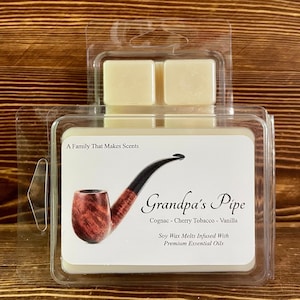Puede incluir: Un recipiente blanco de cera para fundir con una tapa de plástico transparente. El recipiente tiene una etiqueta que dice "Grandpa's Pipe" con el texto "Cognac - Cherry Tobacco - Vanilla" y "Soy Wax Melts Infused With Premium Essential Oils". Una pipa marrón y negra está representada en la etiqueta.