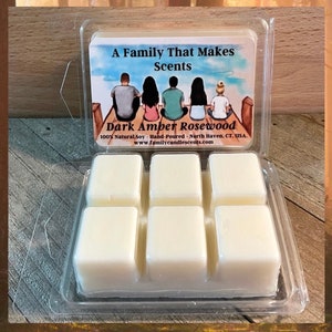 May include: A clear plastic container with six wax melts. The wax melts are white and are arranged in a 2x3 grid. The container has a label that reads "A Family That Makes Scents" and "Dark Amber Rosewood". The label also includes the text "100% Natural Soy Hand-Poured - North Haven, CT. USA." and "www.familycandlescents.com".