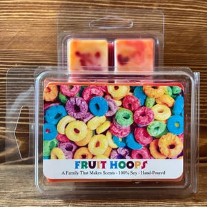 May include: A clear plastic package containing two small orange wax melts with a fruity scent. The wax melts are shaped like squares and have a swirl pattern. The package also includes a larger wax melt with a picture of colorful cereal rings on the front. The text on the front of the package reads "FRUIT HOOPS A Family That Makes Scents - 100% Soy - Hand-Poured".