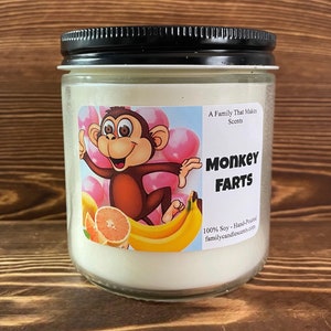 May include: A glass jar candle with a white label featuring a cartoon monkey, bananas, and a grapefruit. The label reads "A Family That Makes Scents", "MONKEY FARTS", "100% Soy - Hand-Poured", and "familycandlescents.com".