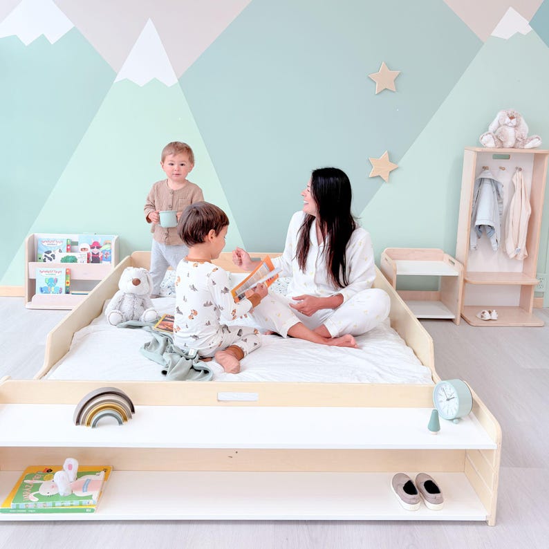 May include: A light wood bed frame with a white mattress, a low shelf, and a matching bookcase. A rainbow wooden toy and a book are on the shelf. A woman and two children are in the room. The wall is painted with a mountain scene.