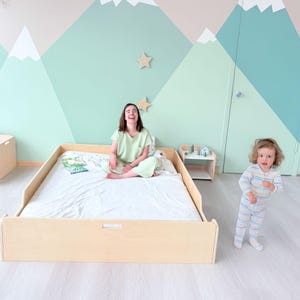 May include: A light wood platform bed with a white sheet. A person in a light green outfit sits on the bed. A child in striped pyjamas stands nearby. The wall has a mountain mural and wooden star decorations.