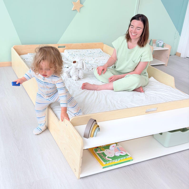 May include: A light wood-framed toddler bed with a white mattress and a lower shelf. A child in striped pajamas plays near the bed, while an adult sits on the bed. A stuffed animal and books are also visible.