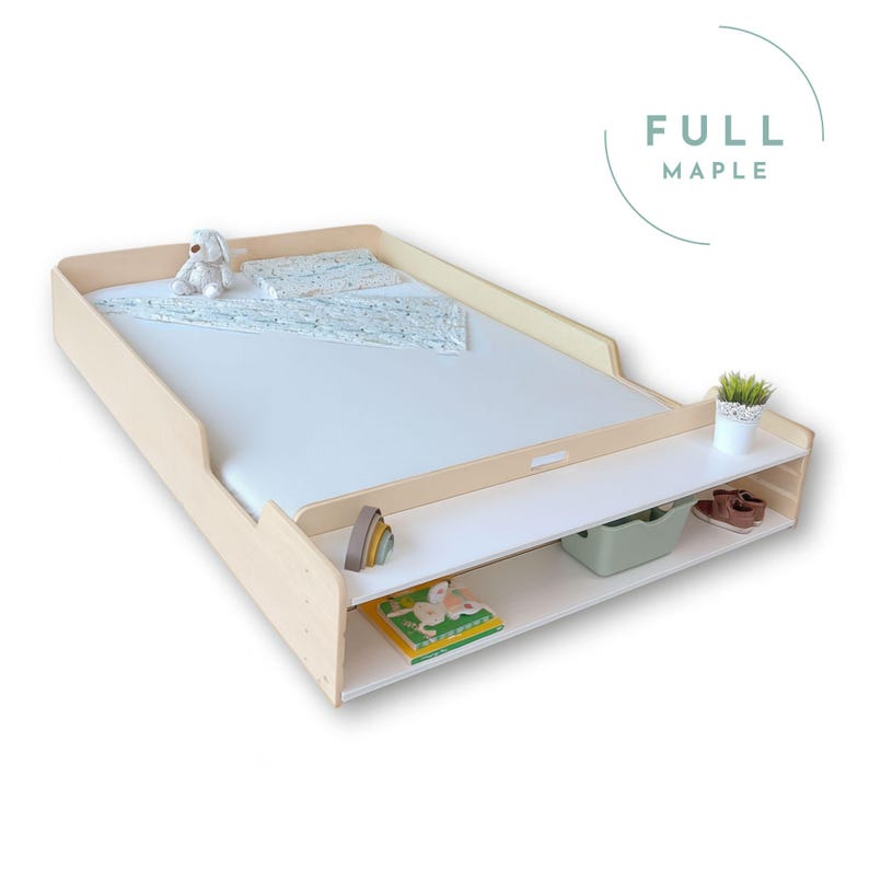 May include: A light wood-toned changing table with a white surface and two shelves. The top surface has a white mattress, a small stuffed animal, and a patterned blanket. The shelves hold a green bin, a book, and other small items. The text "FULL MAPLE" is in the upper right corner.